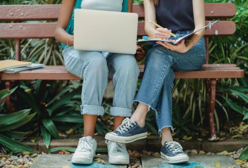 Two women sat on bench with laptop and notebook and pencil
